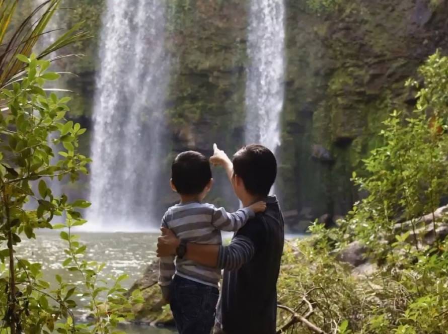 A parent holding a young child, both seen from behind, pointing towards a waterfall