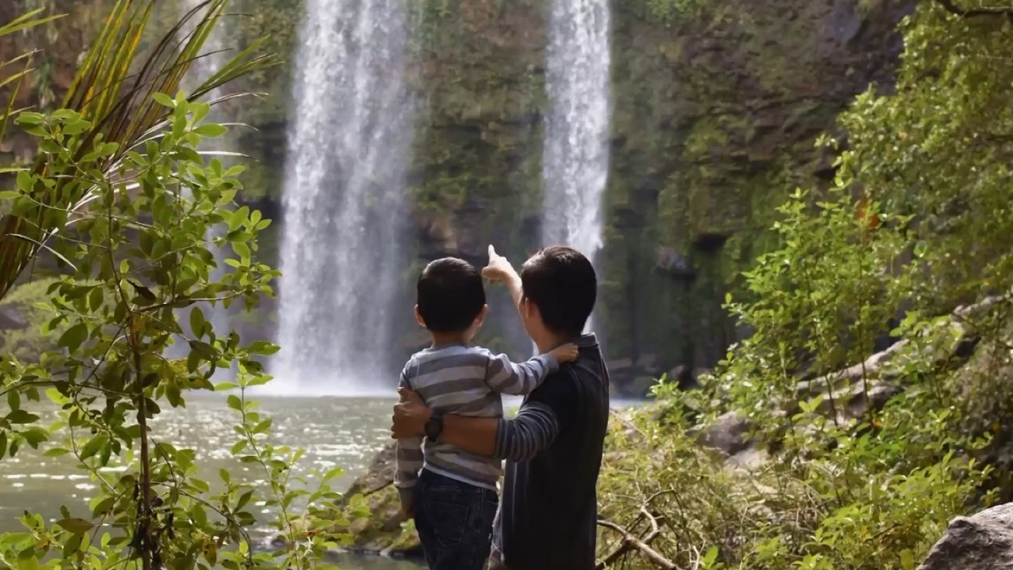 A parent holding a young child, both seen from behind, pointing towards a waterfall